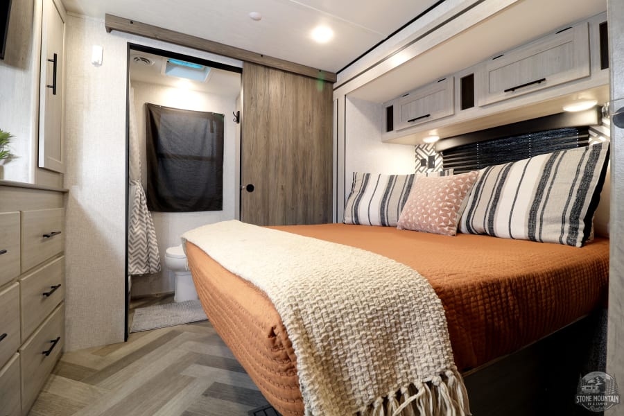 This modern RV rental bedroom features a comfy bed, dresser, overhead cabinets, and a private sliding-door bathroom with a toilet.