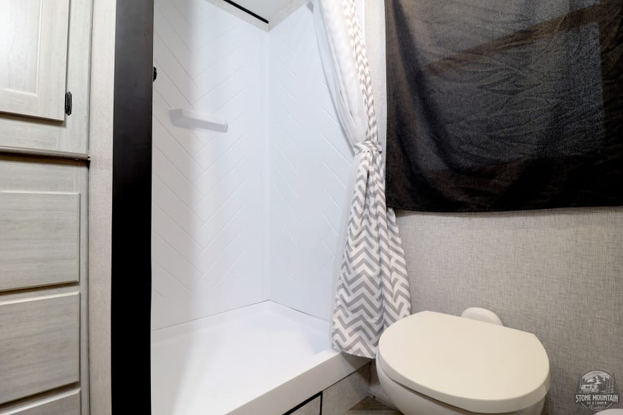 Compact RV rental bathroom featuring a white herringbone shower, tied-back chevron curtain, toilet, and light cabinetry.