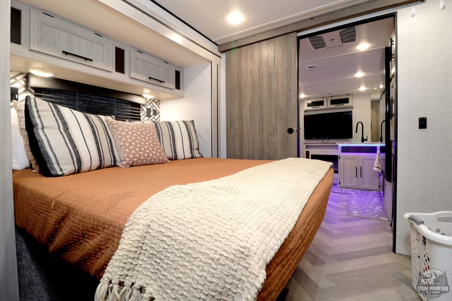 Modern RV rental bedroom with comfortable bed, rust quilt, and neutral accents. Sliding door reveals illuminated living space.