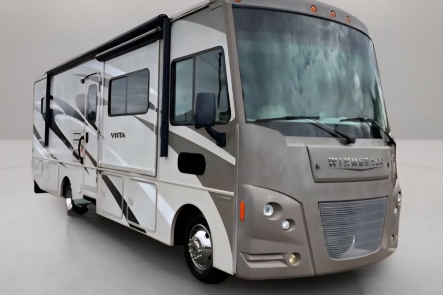 A modern Winnebago Vista rv rental. This tan and white motorhome features an awning, multiple windows, and a detailed front grille.