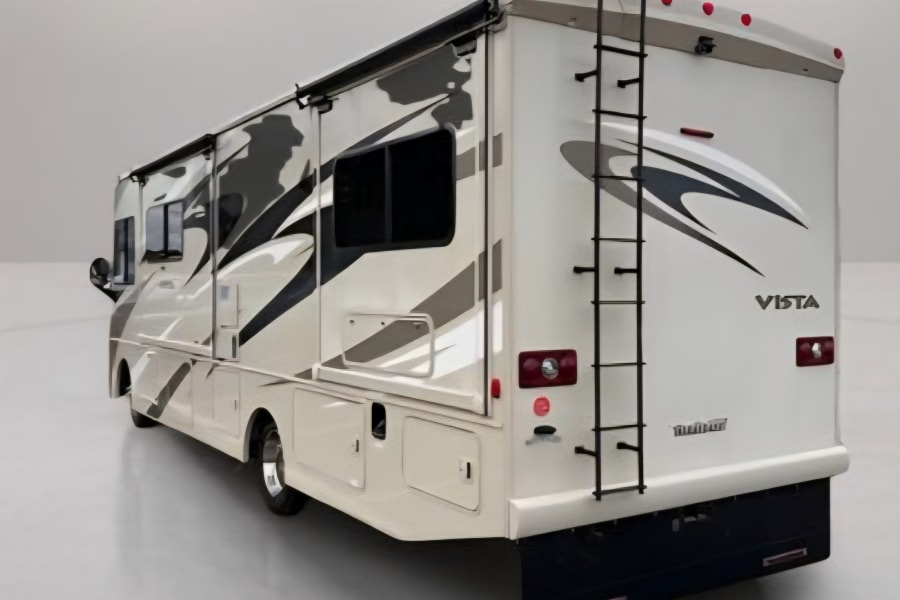 A white Winnebago Vista RV is shown from the rear-right, featuring a ladder and graphics, perfect for an rv rental.