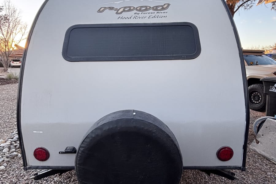 Rear view of an r-pod travel trailer, great for an rv rental. Features a spare tire, window, and tail lights on gravel.