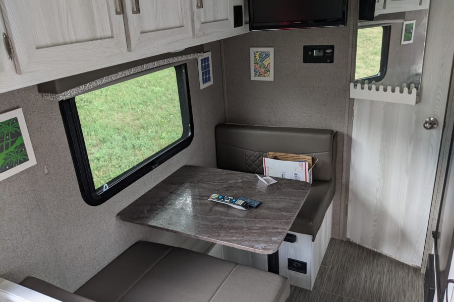 An RV rental dinette interior showing bench seating, a table with remotes, a window view of grass, and overhead cabinets.