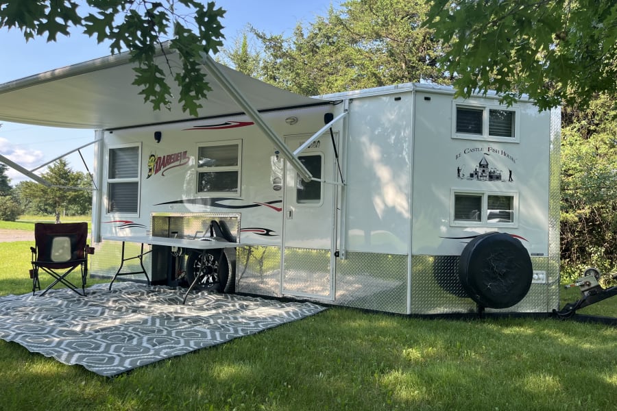 White RV rental, an 'Ice Castle Fish House,' parked on green grass with extended awning, outdoor rug, chair, & table.
