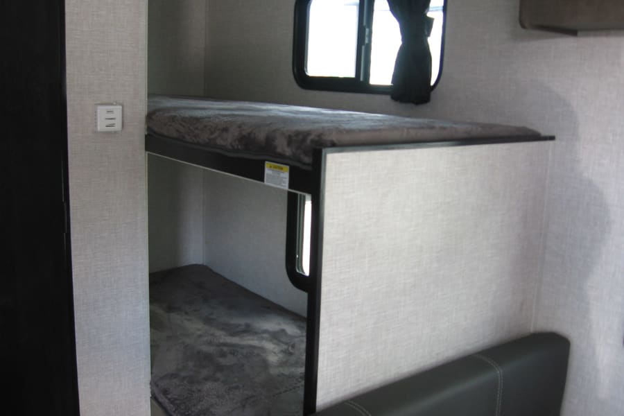 RV rental interior showing stacked bunk beds with dark mattresses. A window, curtain, and thermostat are visible.