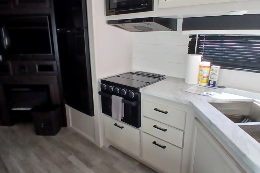Inside an RV rental: a bright kitchen with black appliances, white cabinets, and wood-look flooring, leading to a cozy living area.