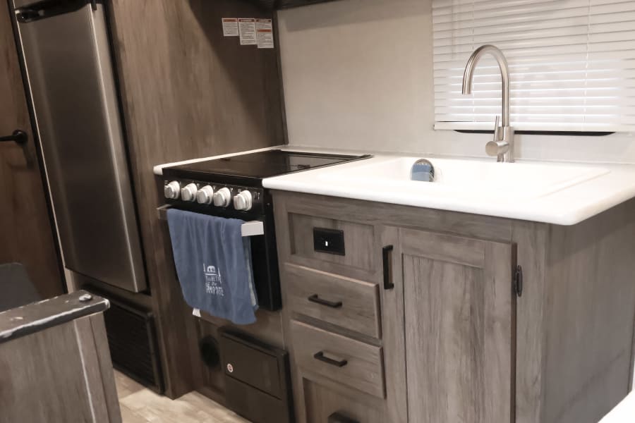 RV rental kitchen: stainless fridge, stove, white sink, wood-grain cabinets, and window blinds. A blue towel hangs on the oven.