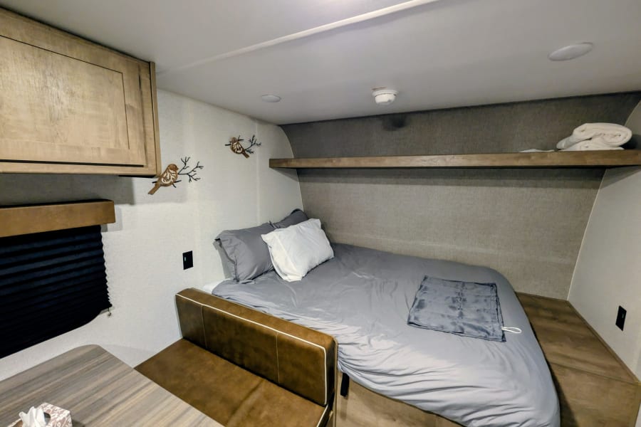 Cozy RV rental interior: A modern bedroom with a gray-linened bed, wooden shelf, and bird decor, plus a compact dining area.