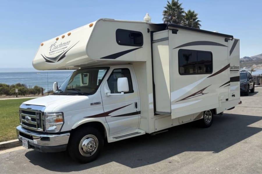 White Coachmen Freelander rv rental, a Class C motorhome with a slide-out, parked by the ocean with palm trees.