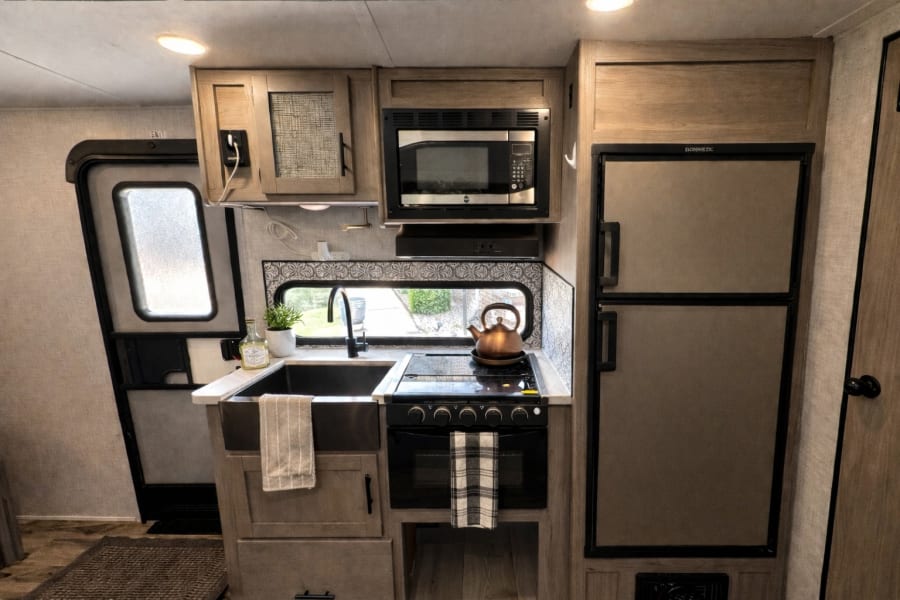 Inside a modern RV rental kitchen: sink, stove, microwave, and fridge. An open doorway with a frosted window is on the left.