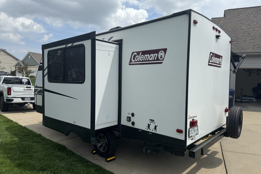 White Coleman travel trailer with a slide-out, likely an RV rental, parked on a driveway next to a GMC truck.