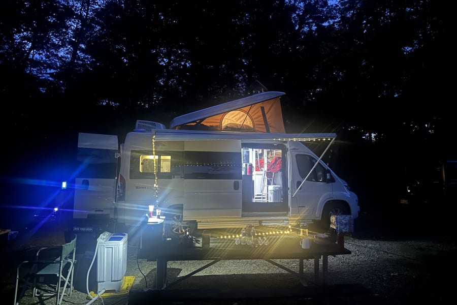 Night camping with an illuminated RV rental, pop-top open, string lights, and a portable washing machine at the campsite.
