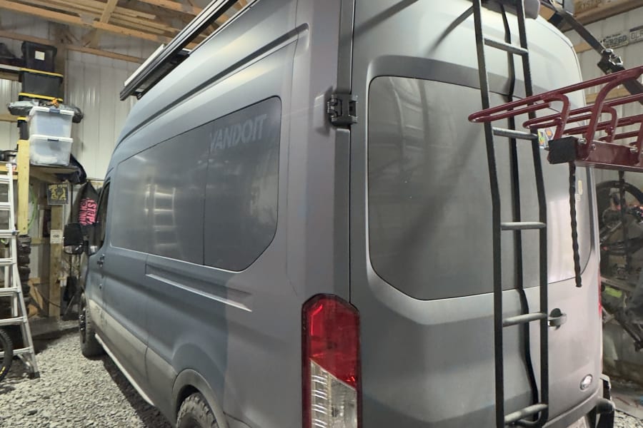 Gray Vandoit camper van (an rv rental option) with roof box, rear ladder, and bike rack, parked on gravel indoors.