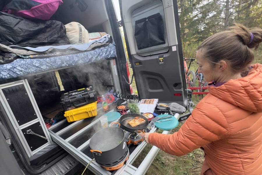 Cooking a fresh meal outside a camper van, possibly an rv rental. A person fries salmon and boils vegetables on portable stoves in a forest setting.