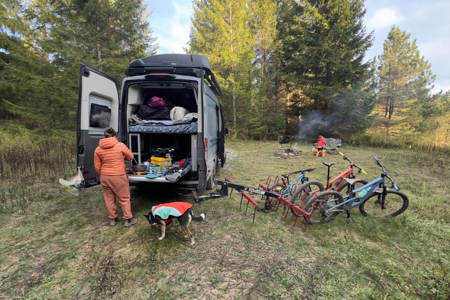 An RV rental provides a cozy camp setup: Cooking by the van, bikes, dog, and a child by the campfire.
