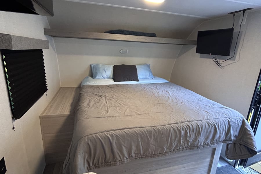 Cozy RV rental bedroom with a comfortable bed, overhead shelf, wall-mounted TV, and a partial view of the kitchen area.