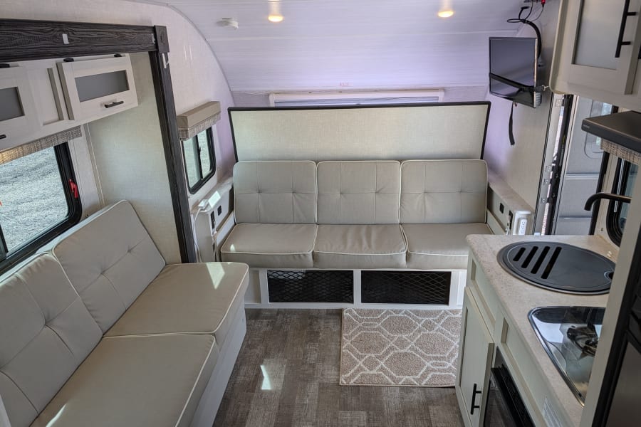 Cozy RV rental interior featuring two light-colored sofas, a compact kitchen with sink/stovetop, and a wall-mounted TV.