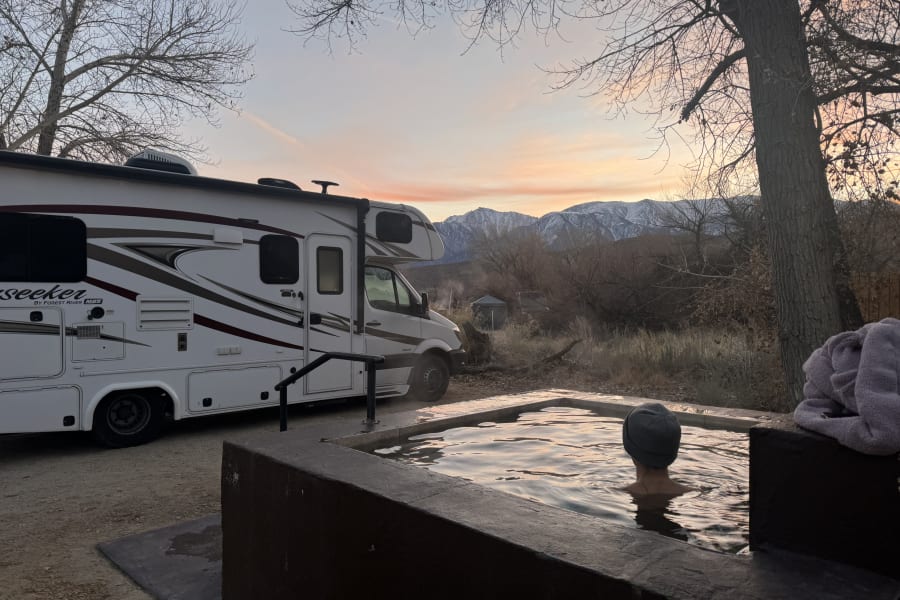 Person soaking in a hot spring with a Forest River Seeker RV rental parked nearby. Snowy mountains and sunset sky in the distance.