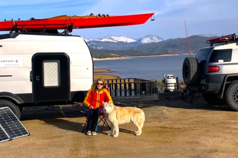 Woman and dog with a Beyond Grid Campers rv rental, SUV, kayak, solar panels, and generator by a scenic lake & mountains.
