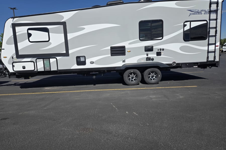 Modern grey travel trailer RV rental, featuring unique white and grey decals, parked on asphalt under a sunny sky.