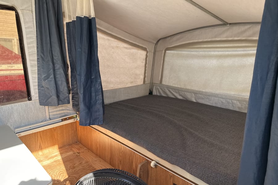 RV rental pop-up camper interior: a bed with gray mattress, blue curtains, and fan, ready for camping.