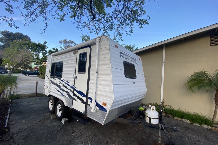 White Carson Rebel rv rental with blue accents, parked on an asphalt driveway beside a house under a clear blue sky.