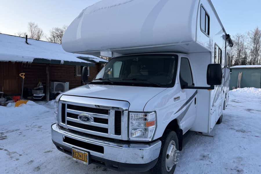 White Class C rv rental, a Ford E-450, covered in snow, parked near a cabin in a winter landscape.