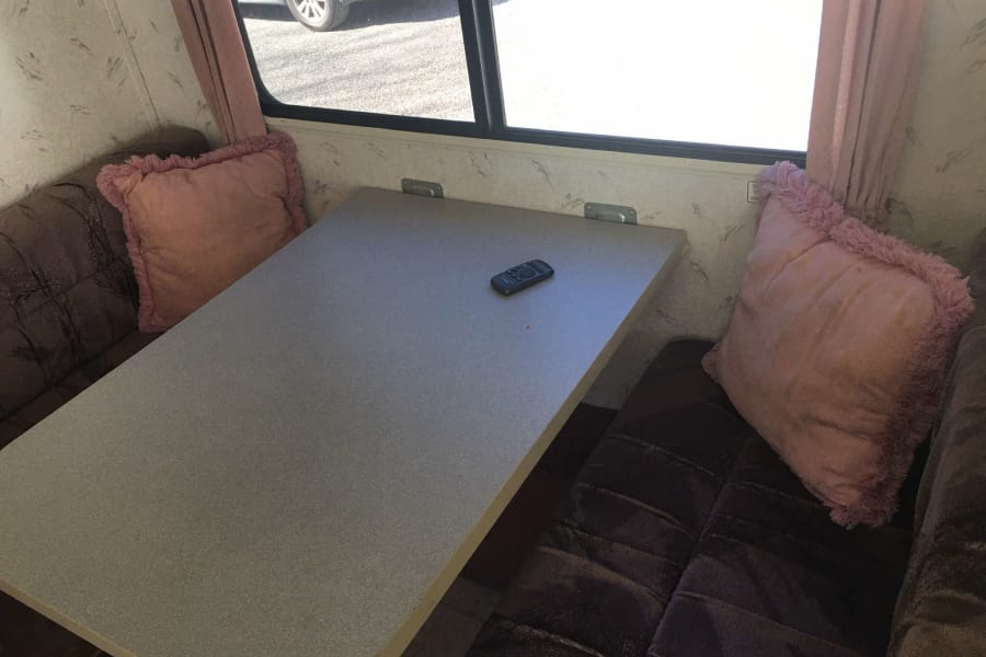 RV rental dining area: a light grey table with a remote, flanked by dark brown benches with pink pillows by a window.