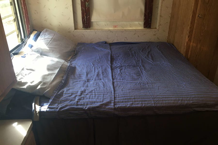 Inside an RV rental: a bedroom with a blue striped bed, pillows, window on the left, and an overhead EXIT sign.