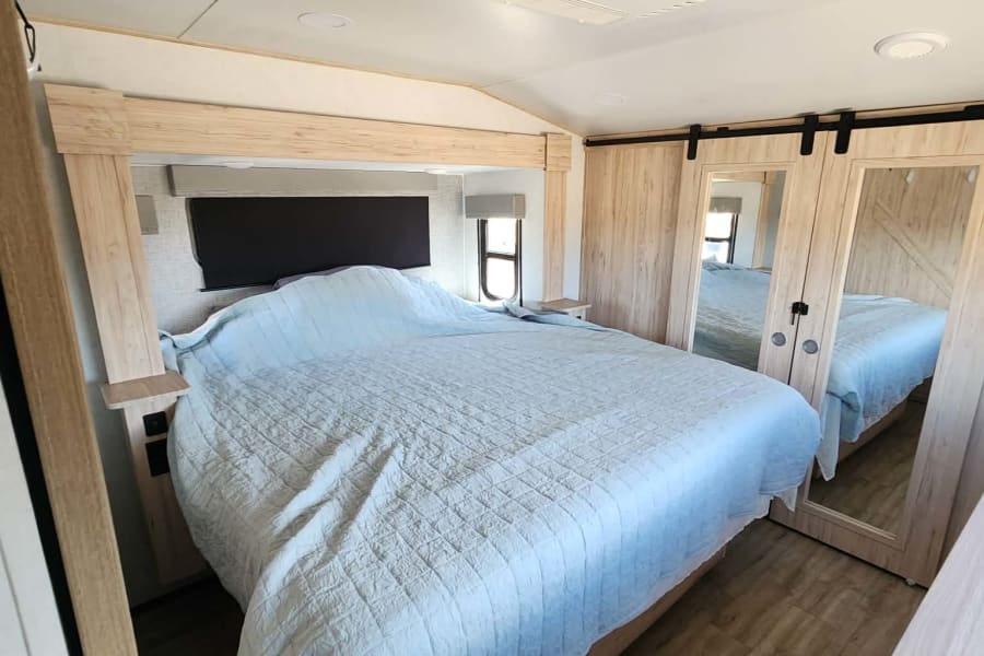 Modern RV rental bedroom featuring a large bed, light wood headboard, and mirrored sliding closet doors.