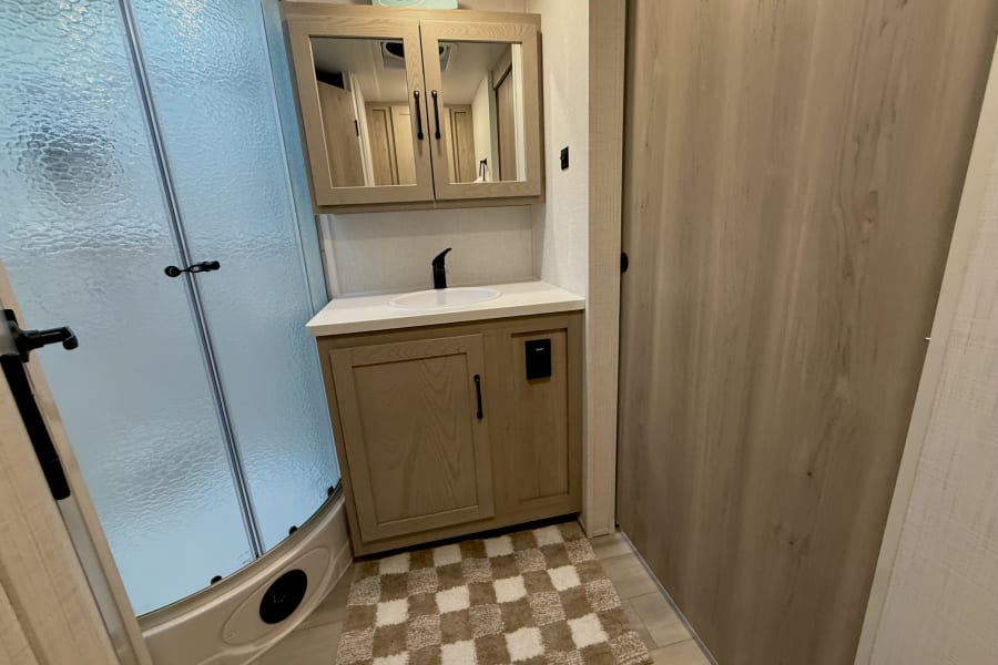 This RV rental bathroom features a compact shower, light wood vanity with a sink, and a cozy checkered rug.