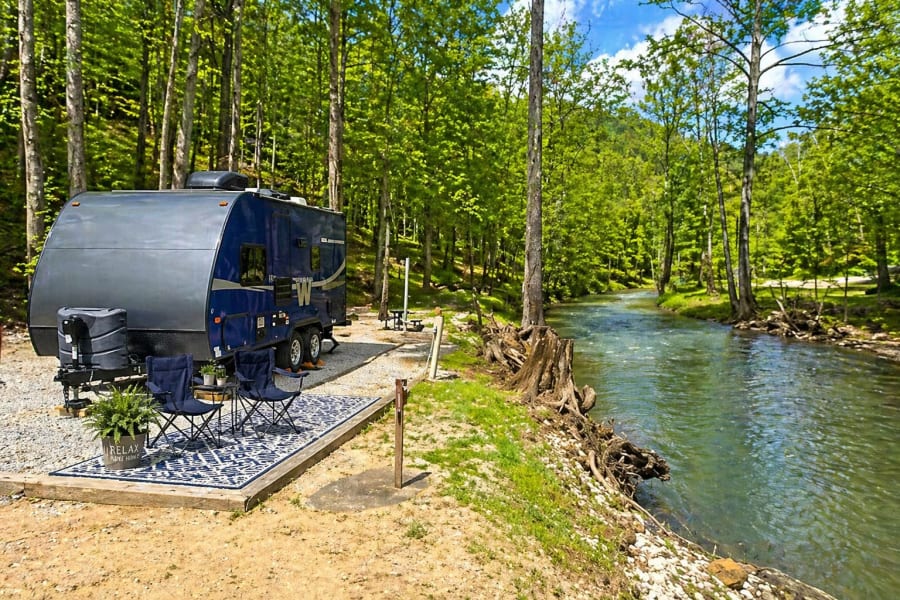 This scenic RV rental site, complete with outdoor seating, is nestled by a clear river in a lush, green forest, ideal for camping.