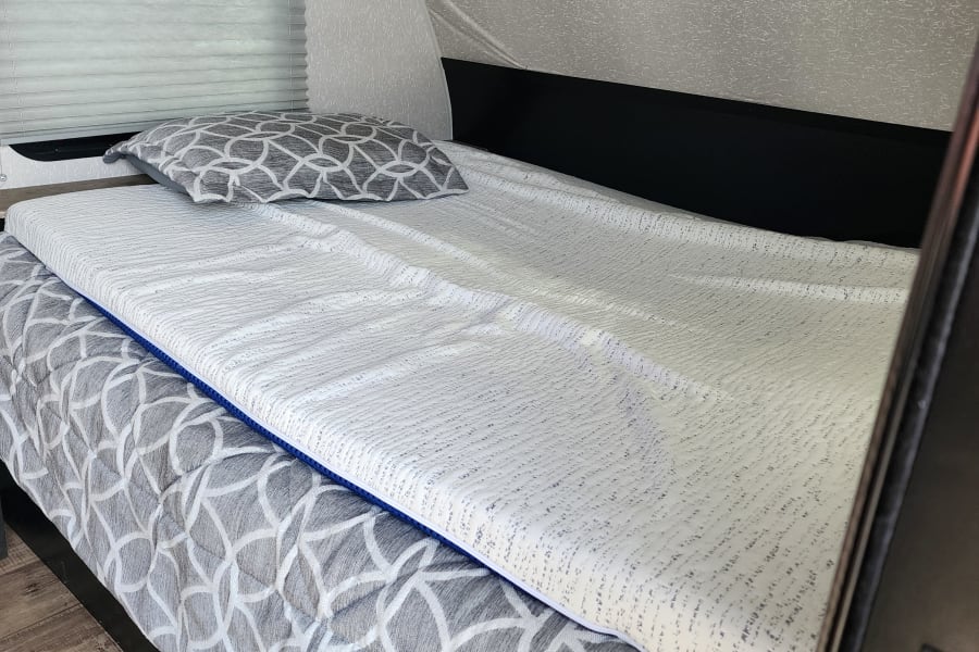 Cozy rv rental interior with a bed made up with gray patterned bedding, a white textured mattress topper, and window blinds.