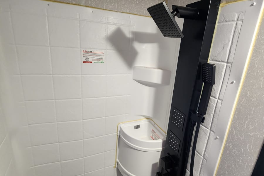 A compact RV rental shower with white, tile-patterned walls, a modern black shower panel, and a corner sink.