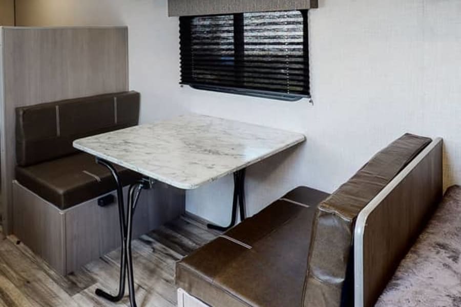 RV rental dinette with brown upholstered benches, a white marble-look table, and a window with blinds for comfortable travel.