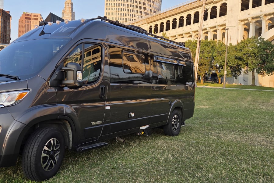 Dark gray Winnebago camper van, perfect for an rv rental, parked on green grass. City buildings & arched bridge in background.