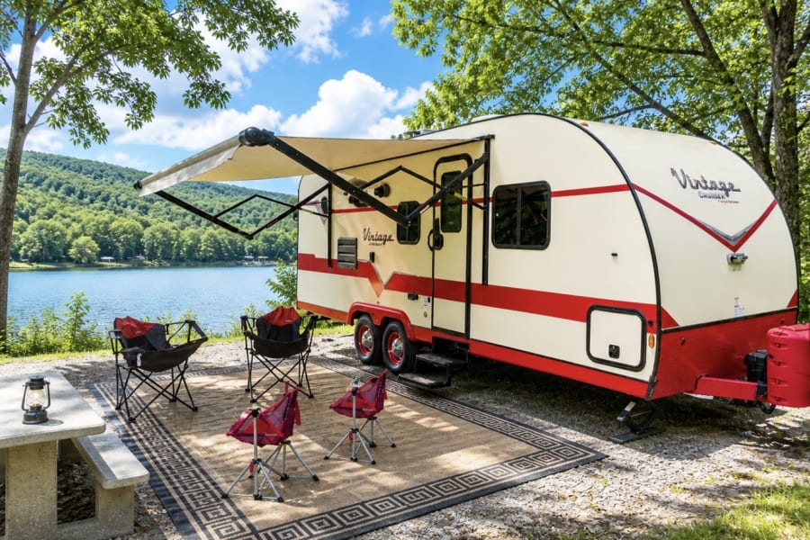 Scenic rv rental at a lakeside campsite. Vintage camper with awning, outdoor rug & chairs, surrounded by trees & mountains.
