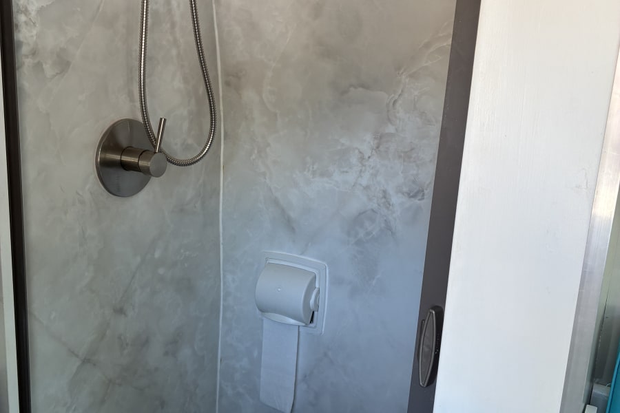 RV rental compact bathroom with shower, toilet, toilet paper, grey marble walls, and a teal floor mat.