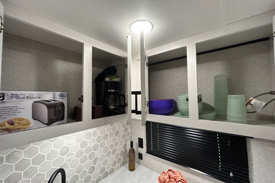 RV rental kitchen: open cabinets reveal a toaster, coffee maker, dishes, and backsplash above a clean counter.