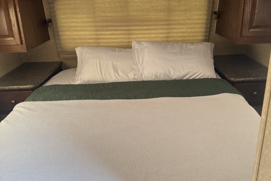 Cozy RV rental bedroom features a bed with white pillows, green quilted blanket, overhead wood cabinets, and window blinds.