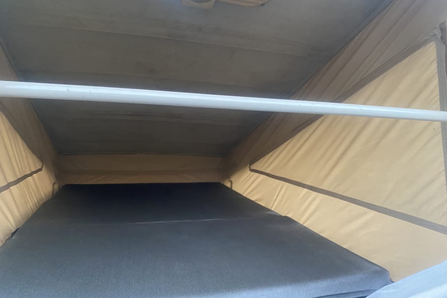 RV rental pop-top camper interior: elevated sleeping area with a gray textured mattress, tan fabric walls, and a white support bar.