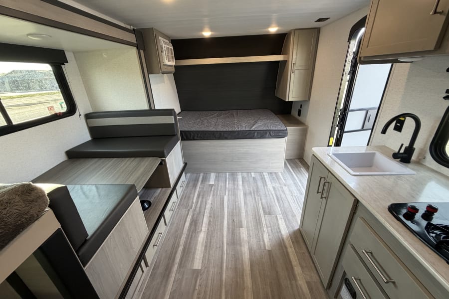Inside a modern rv rental, featuring a bed, dinette seating, and a kitchen area with sink and stovetop. Note the wood-look flooring.