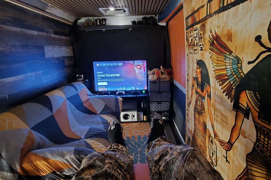 Cozy RV rental interior with a person relaxing on a geometric couch, watching TV. Features unique Egyptian decor.