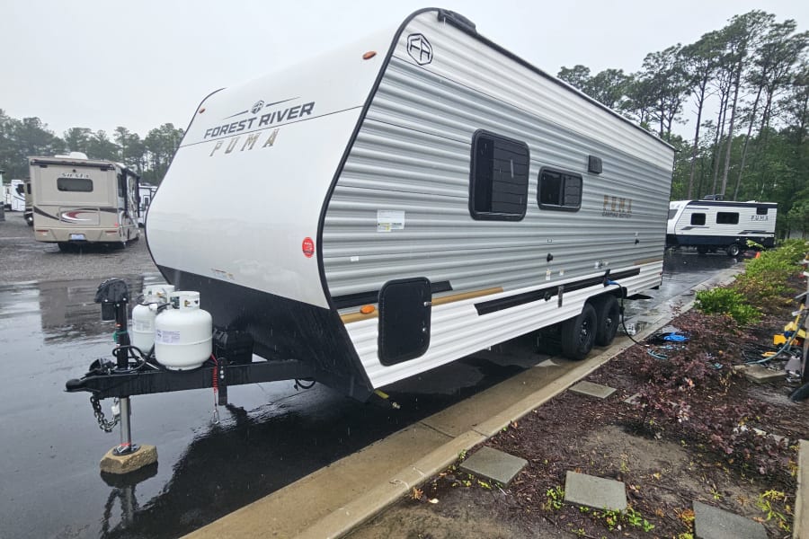 A Forest River Puma travel trailer, ready for RV rental, shown parked on a rainy day with puddles reflecting the scene.