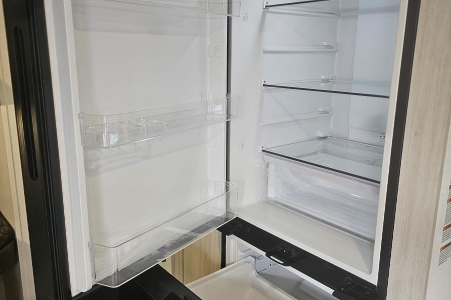 Clean, empty interior of an RV rental refrigerator, featuring clear shelves, door bins, and a partially open pull-out freezer drawer.