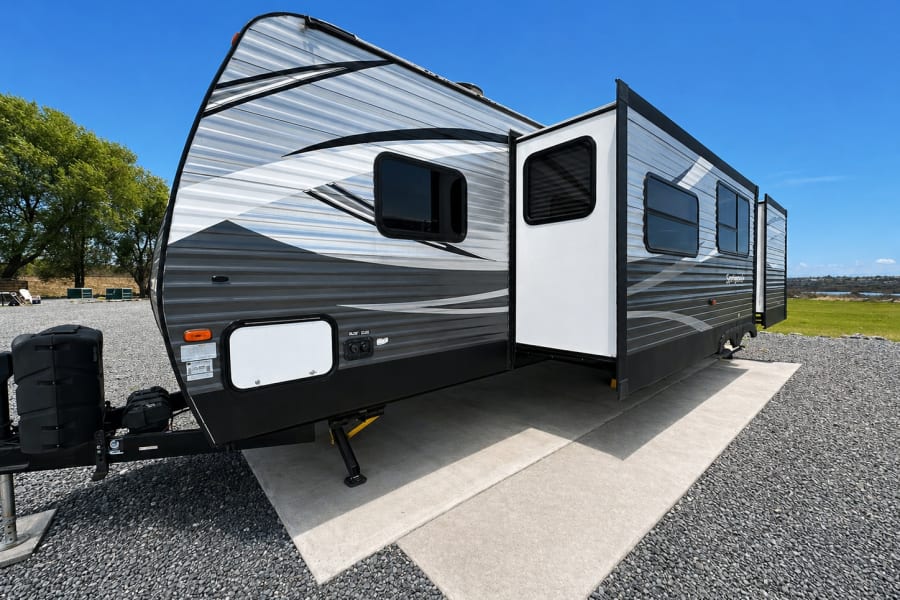 Gray travel trailer RV rental with extended slide-out, parked on a gravel site under a clear blue sky.