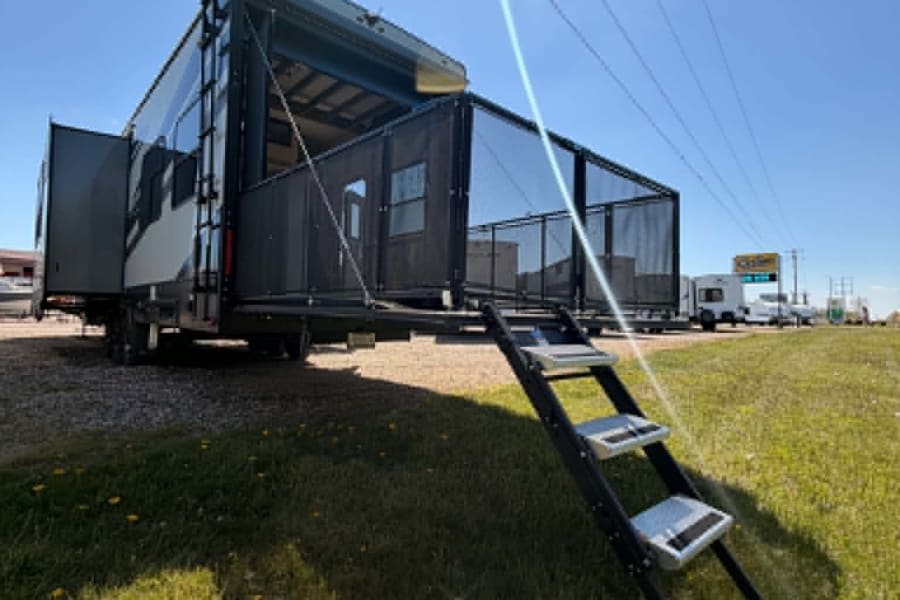 This RV rental is a toy hauler with its rear patio deck and steps deployed, plus a side extension, ready for outdoor adventures.