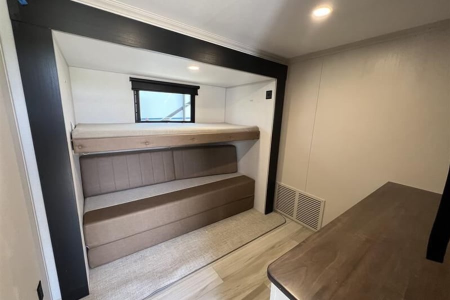 This RV rental bunkhouse offers a cozy interior with a top bunk, convertible sofa bed below, window, and wood-look flooring.