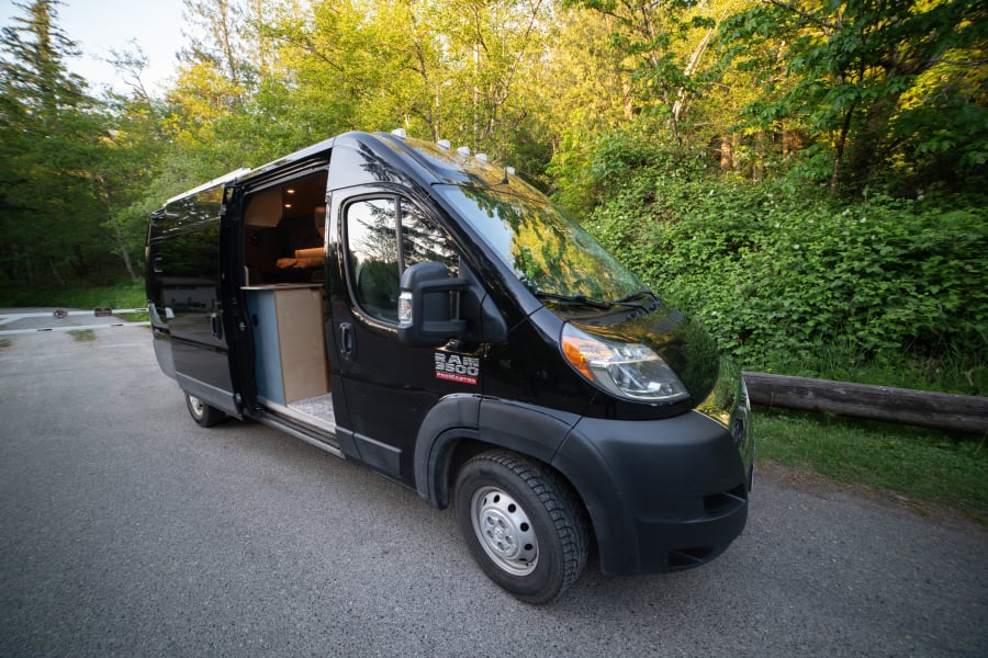 Black RV rental camper van parked on an asphalt road by a green forest. Its open side door reveals a cozy wooden interior.