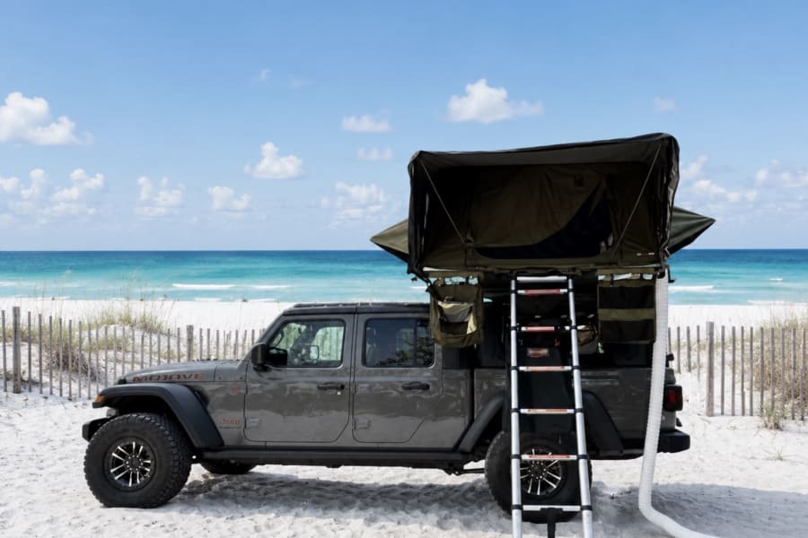 Experience beach camping! A gray Jeep Gladiator with rooftop tent & ladder parked on a white sand beach by the ocean. Ideal for an rv rental.